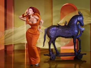Doja Cat: Gone with the Wind by David LaChapelle Doja Cat in an orange sculpted gown bends forward beside a dark horse sculpture on wheels; left hand raised to her forehead; glossy gold floor; orange and yellow arches with a flowing yellow fabric ribbon; poised, mid-gesture mood.