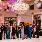Group of young men and women dressed formally while standing in a ball room in front of a stage.